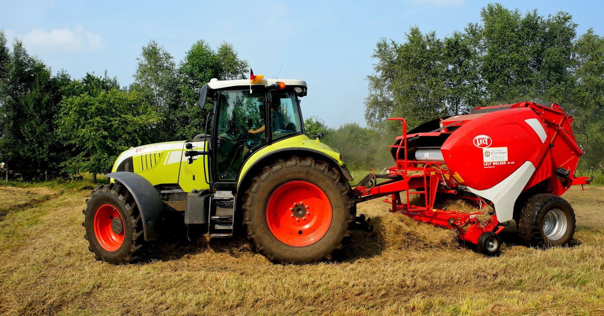 Green tractor and hay baler working on a farm field with trees in the background.