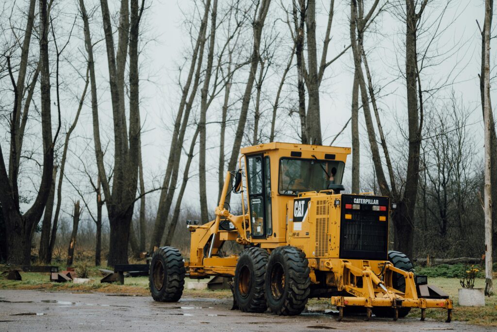 A yellow Caterpillar grader parked in a forest clearing during a cloudy day.